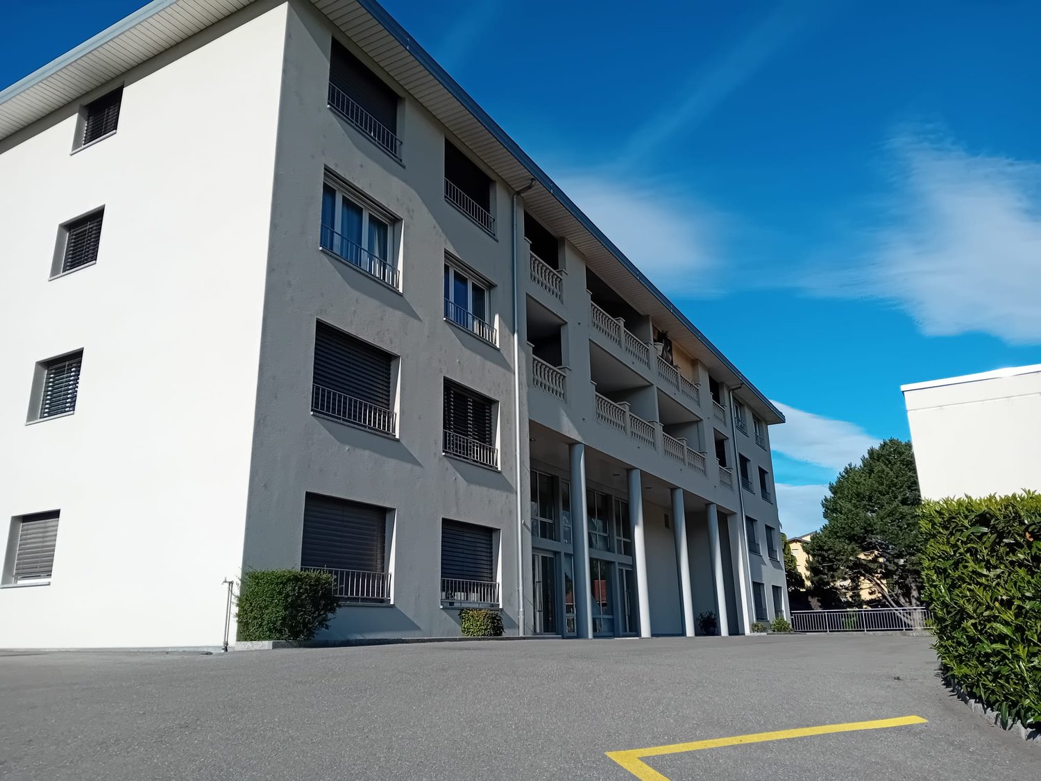 Modern white building, with multiple balconies, glass windows, a fenced area in the front.