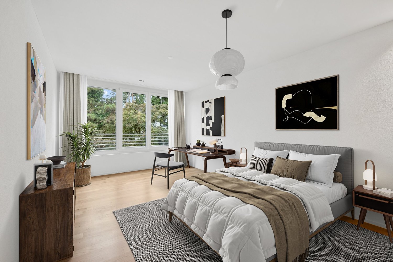 Spacious bedroom with large windows overlooking a wooded area, modern decor with a gray upholstered bed, wooden nightstands, and abstract artwork on the walls. The room also features a desk area and a pendant light fixture.