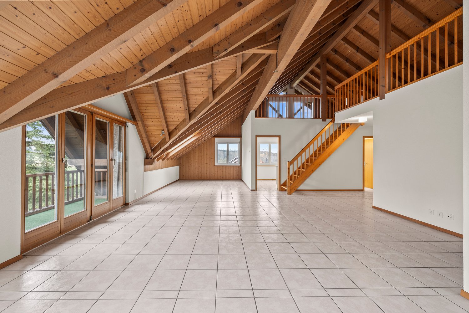 Spacious open-concept living area with vaulted wood-beam ceiling, large windows and sliding glass doors leading to a balcony or terrace, hardwood floors, and a staircase leading to an upper level.