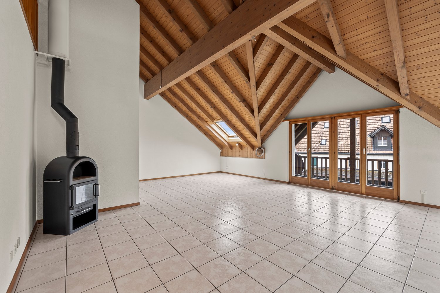 Spacious open-plan living area with vaulted wooden ceiling, large windows providing natural light, tiled flooring, and a fireplace in the corner.