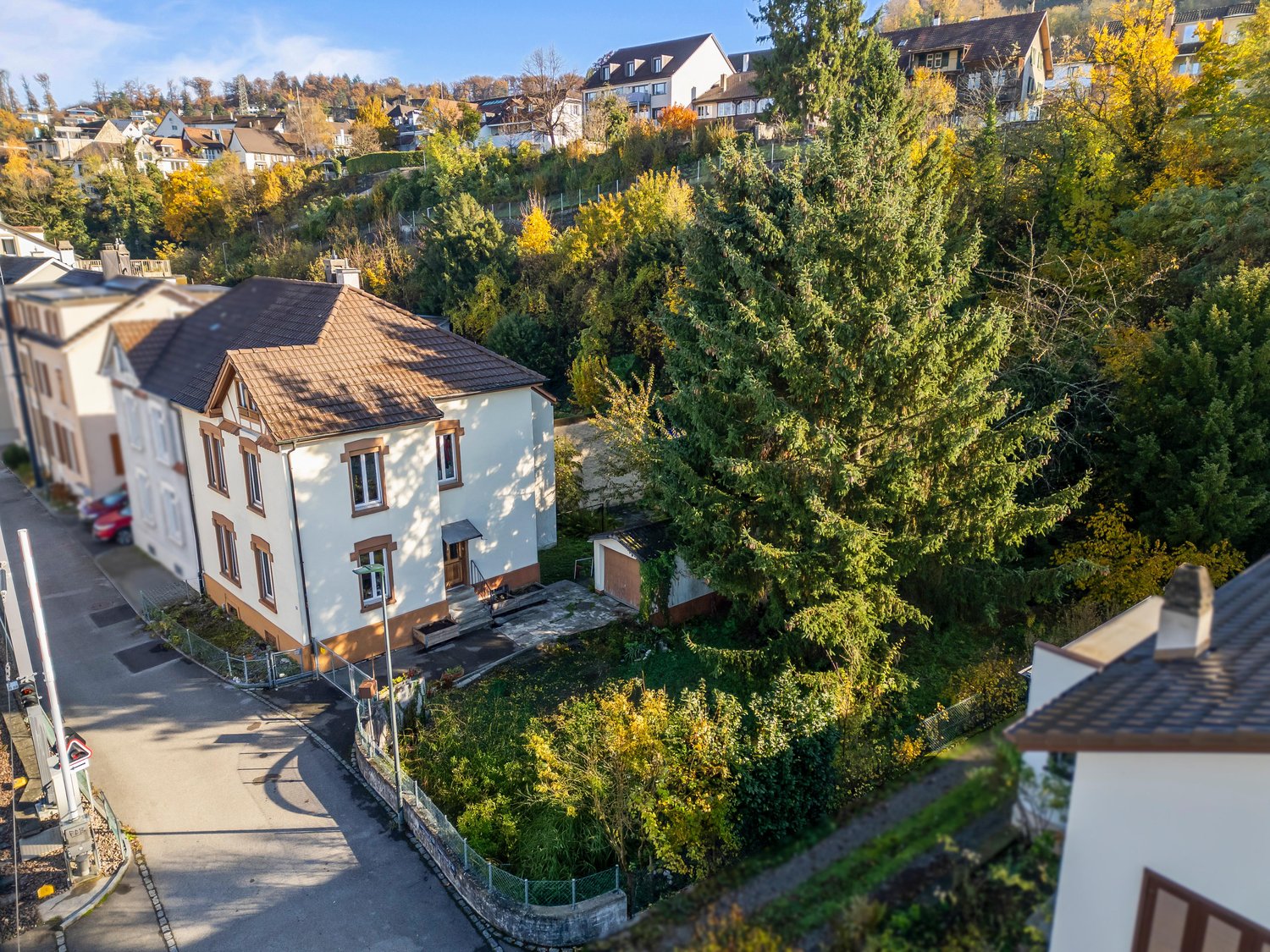 Two-story house with brown roof, white walls, large garden, driveway, several trees, fenced garden, neighboring houses in the background.