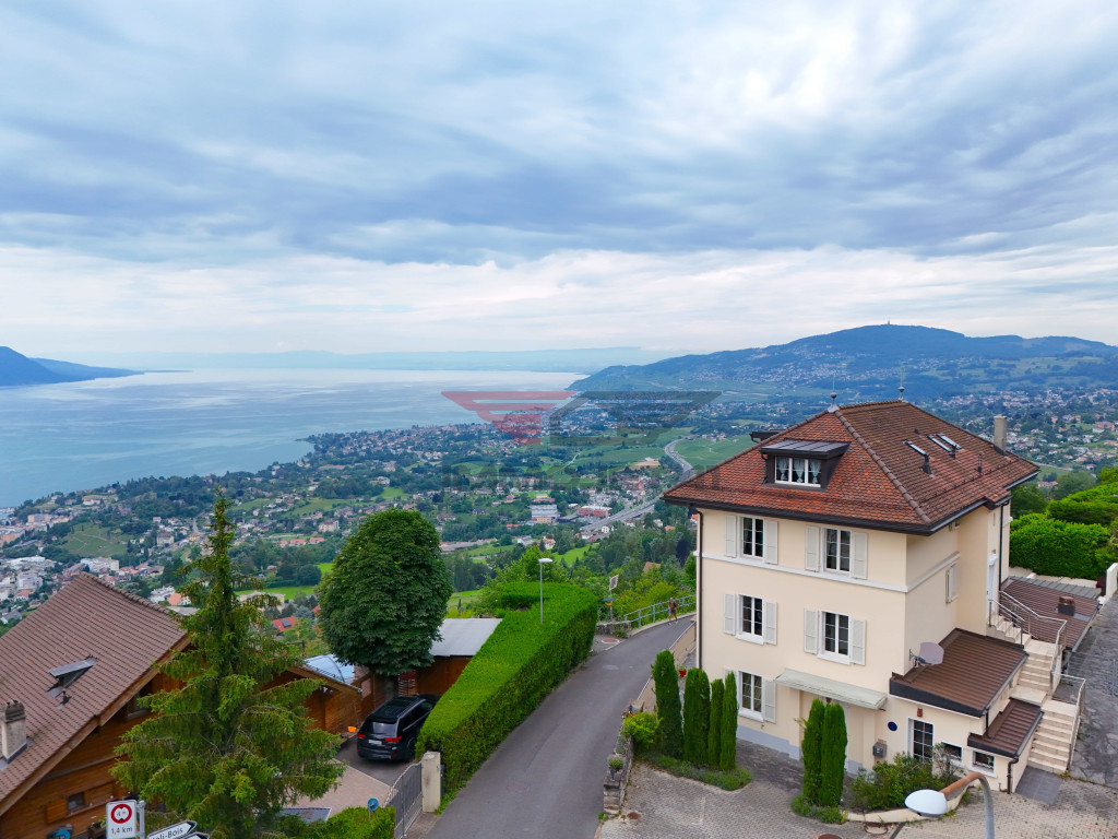 A large multi-story house with a red tile roof, surrounded by lush greenery and overlooking a scenic landscape with a lake, mountains, and a city in the distance.