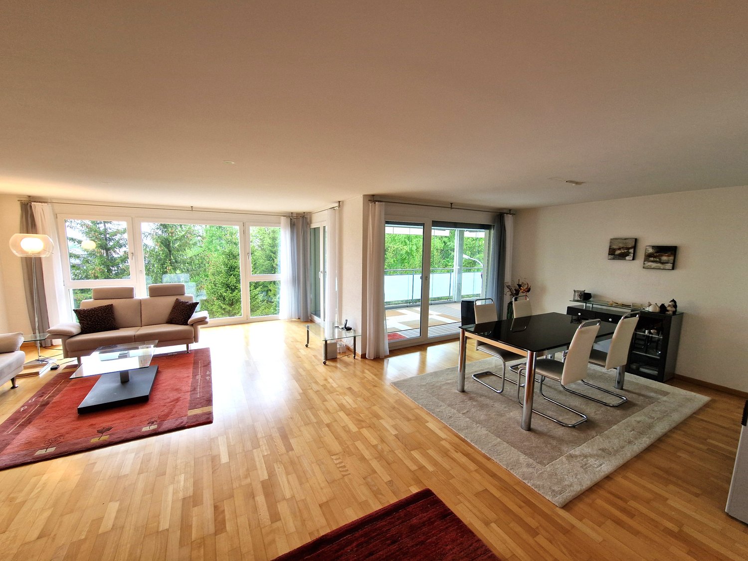 Spacious open-plan living and dining area with large windows overlooking a balcony or terrace. The room features hardwood floors, a brown leather sofa, a coffee table, and a dining table with chairs. There is also a media console and shelving unit against