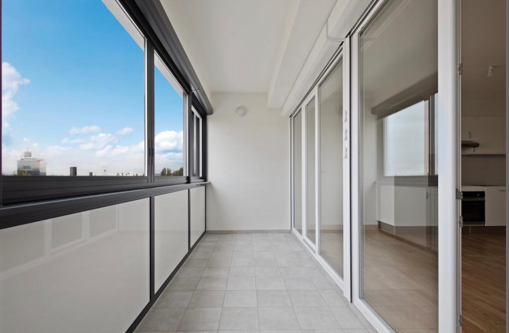 Spacious balcony with large windows, providing a panoramic view of the city skyline. The balcony has a tiled floor and is connected to the interior living space through sliding glass doors.