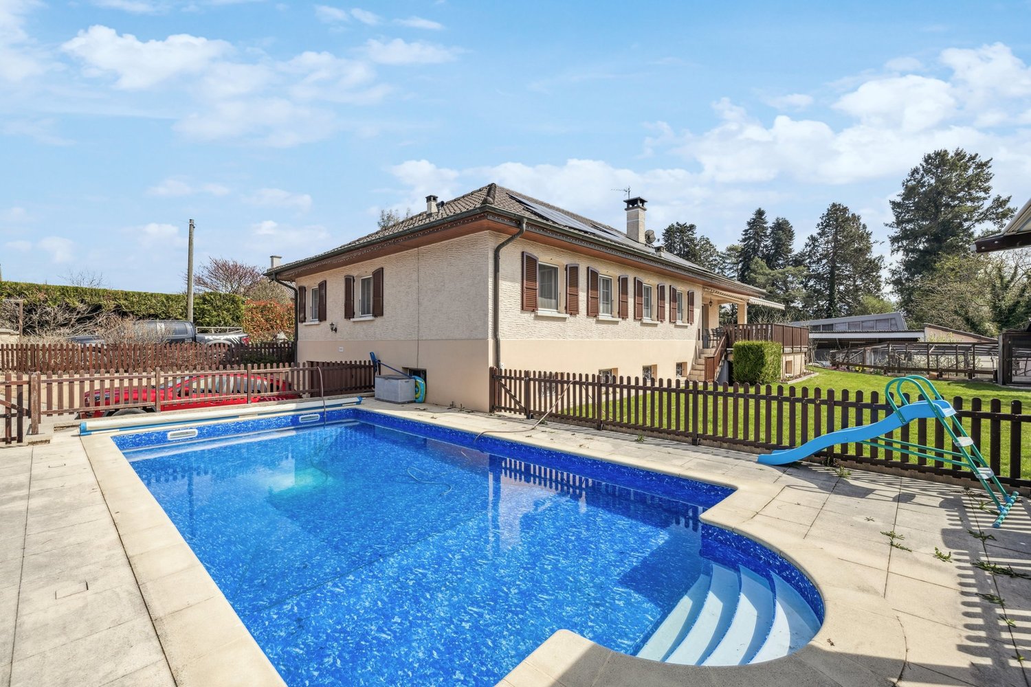 Two-story house with a tiled roof, surrounded by a wooden fence. The house has a swimming pool in the backyard with a ladder and other pool accessories. The property also has a garden and some trees around it.