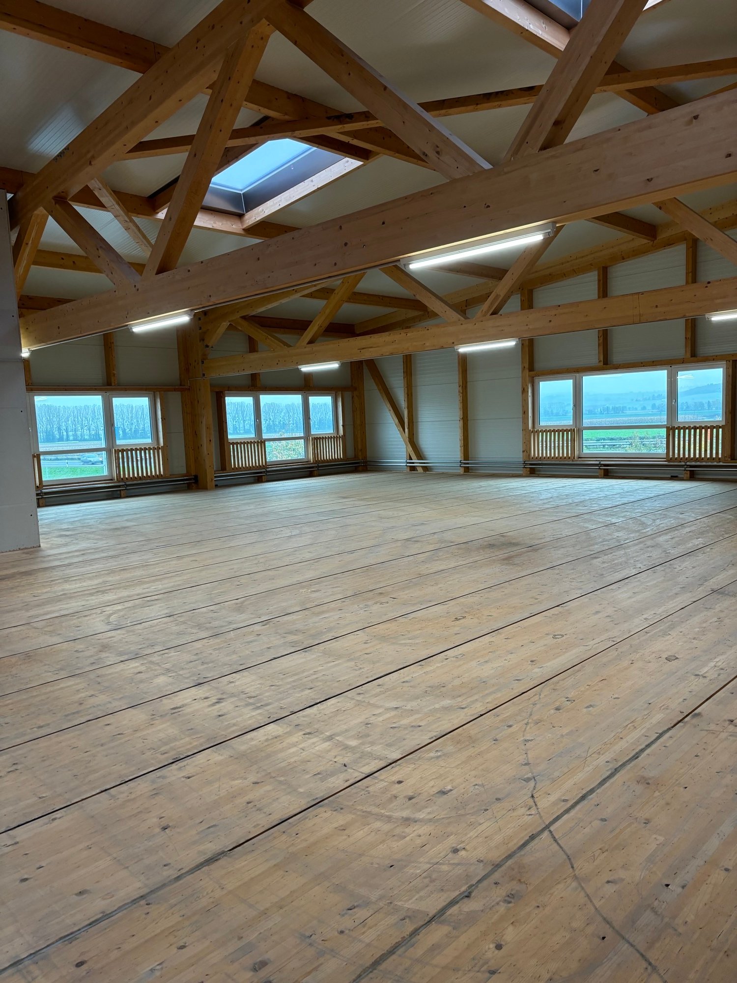 Large open space with wooden beams and trusses, hardwood flooring, and large windows providing natural light and a view of the surrounding landscape.