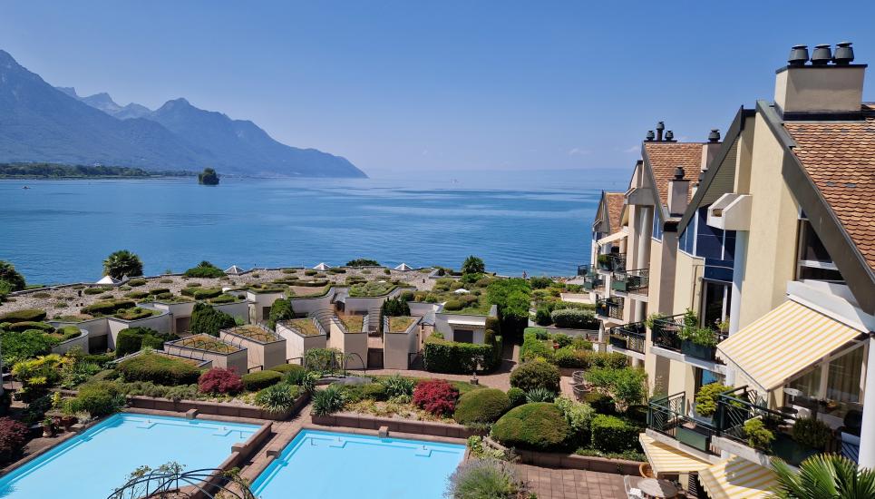 This image shows a scenic apartment complex overlooking a beautiful lake surrounded by mountains. The complex features a series of terraced buildings with balconies and patios, as well as a large swimming pool in the foreground. The buildings have a Medit