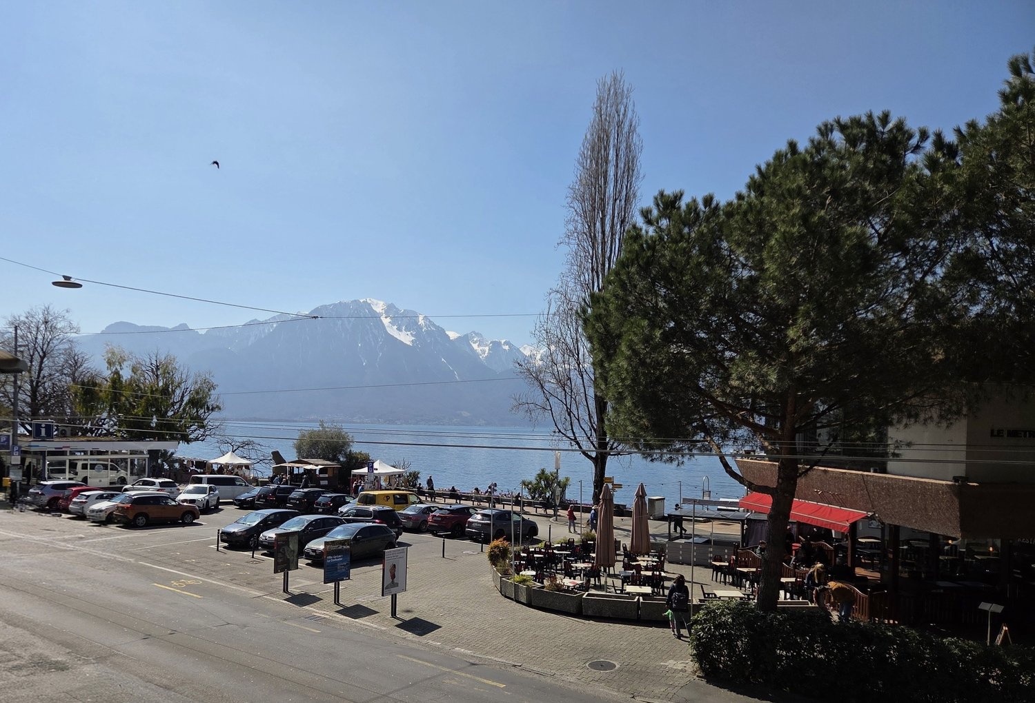 Outdoor parking lot with many cars, a lake, mountains, and a restaurant with outdoor seating.