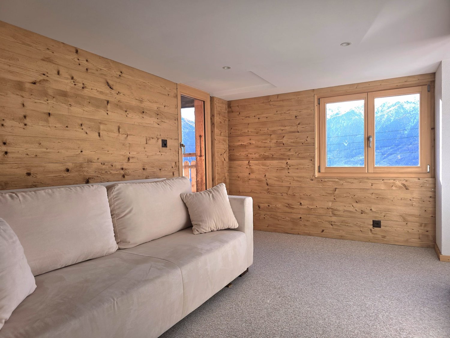 Wooden walls, beige couch, carpeted floor, windows with view of mountains