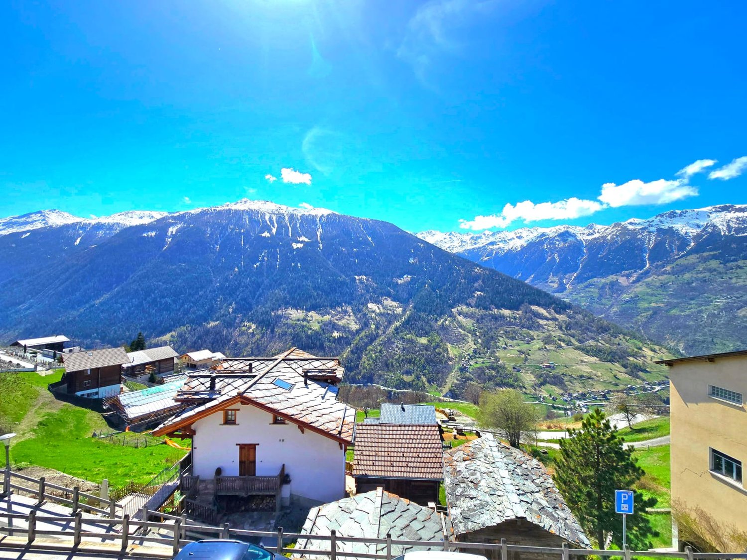 Mountain village with several houses, snow-capped mountains, green fields, clear blue sky