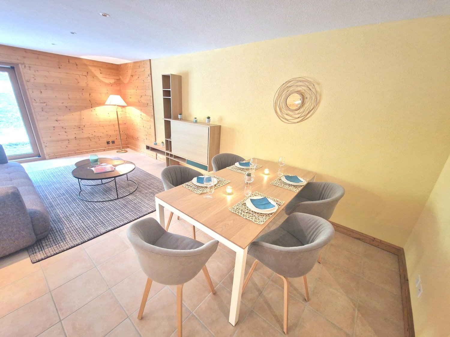 The image shows a combined living room and dining area. The living room has a wooden accent wall, a round decorative wall hanging, and a coffee table. The dining area has a wooden table with four gray upholstered chairs, and a sideboard unit. The room has