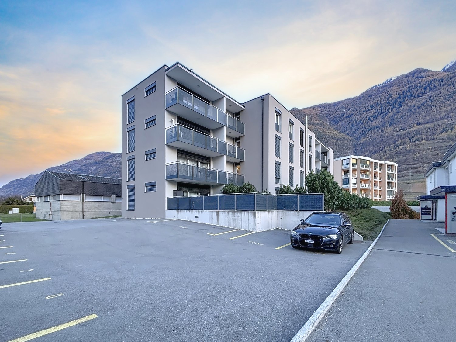 Modern apartment building with multiple balconies, situated at a lower level with a view of the mountains