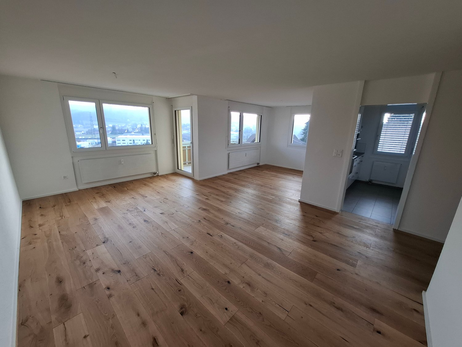 Spacious open-plan living area with large windows, hardwood flooring, and a view of the city outside. The room appears to be a living room or combined living/dining area.