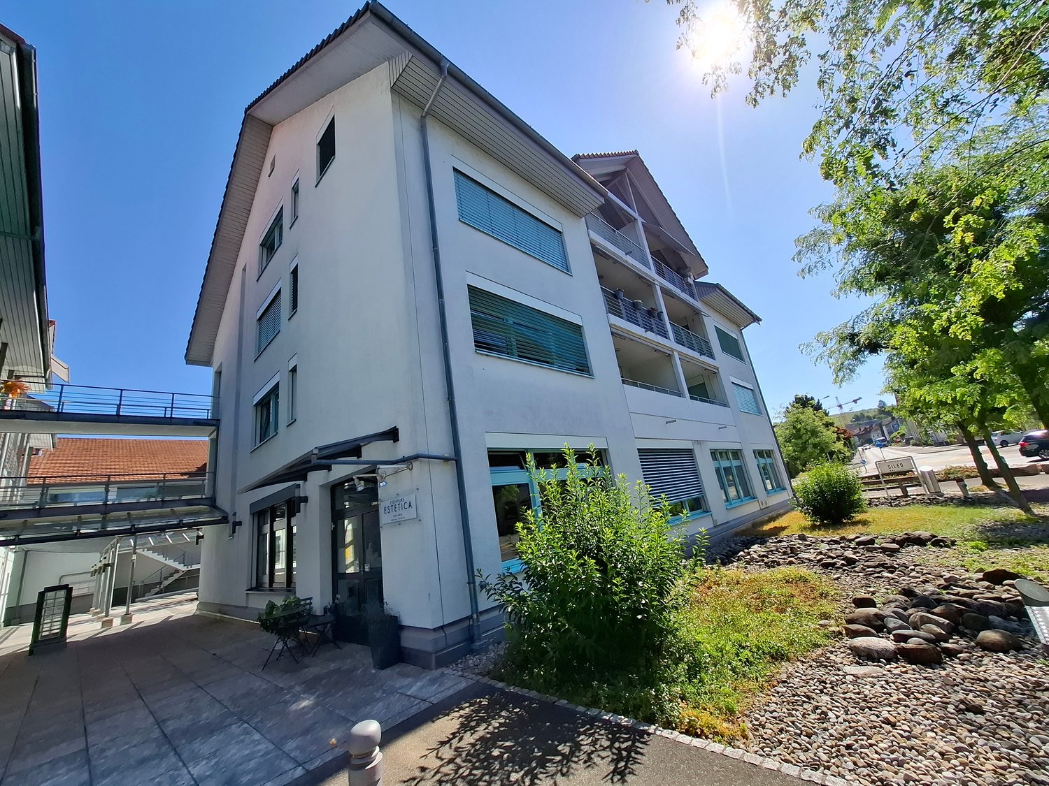 Multi-storey building, white walls, blue window frames, balconies, walkway connecting buildings, plants, gravel, walkway