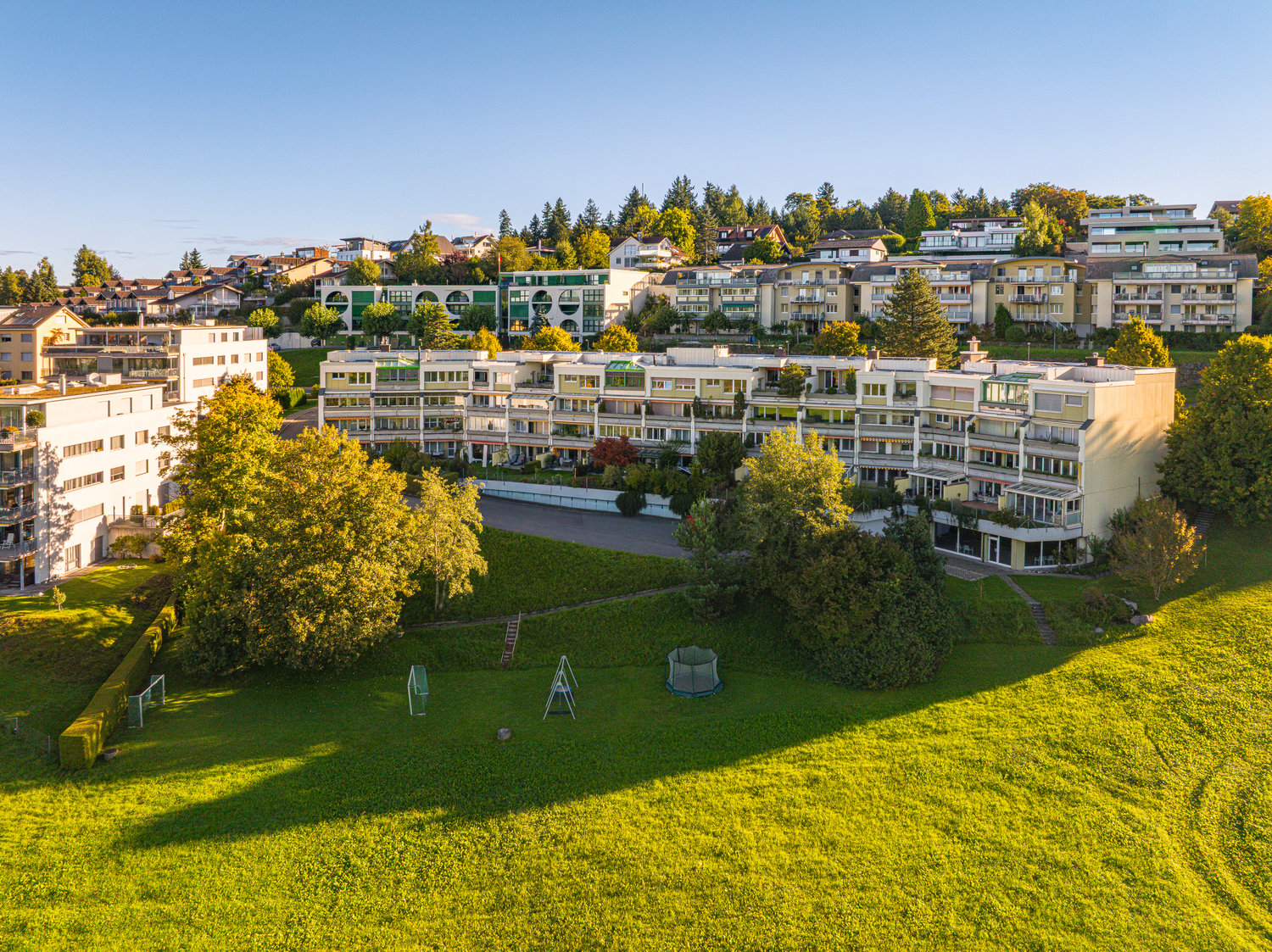 An apartment building complex, several apartment buildings in a row, grassy field with playground equipment, surrounded by trees and bushes.