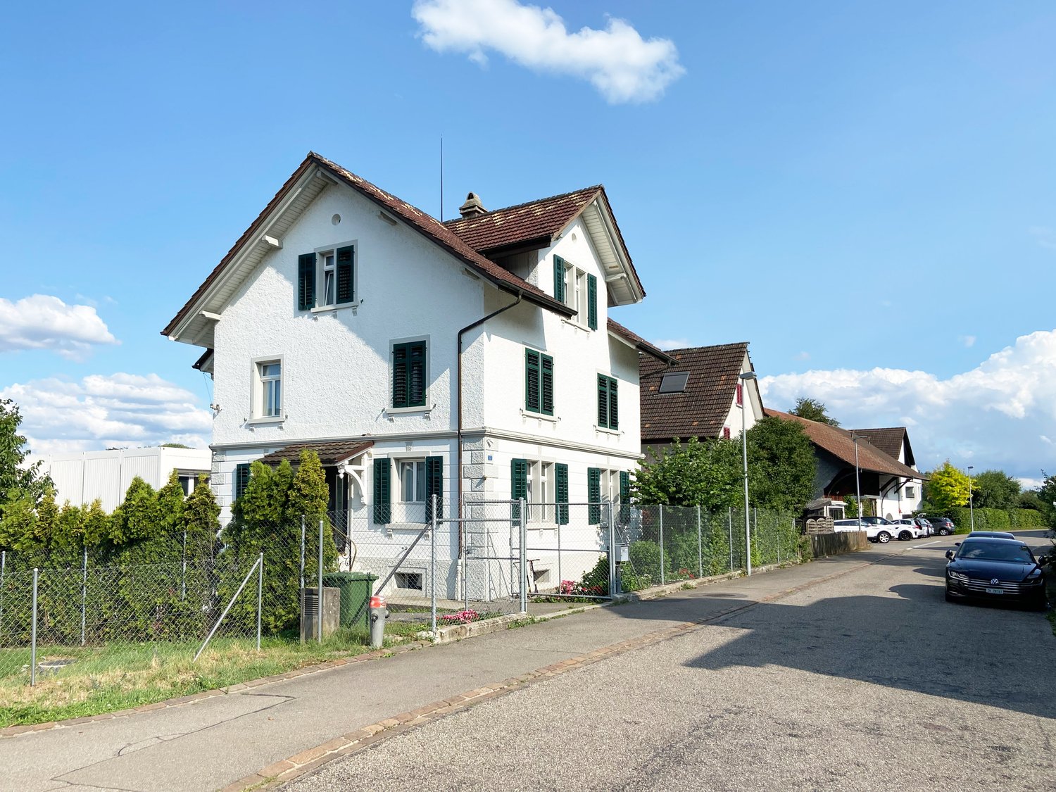 3 story white house, brown roof, green shutters, brick ground, fence, cars parked