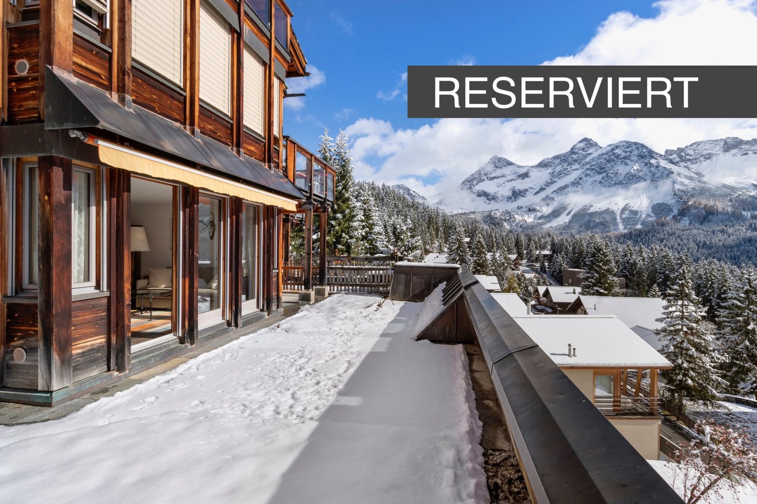 Chalet style building with balcony, snow covered ground, mountain views, multiple houses in the background.