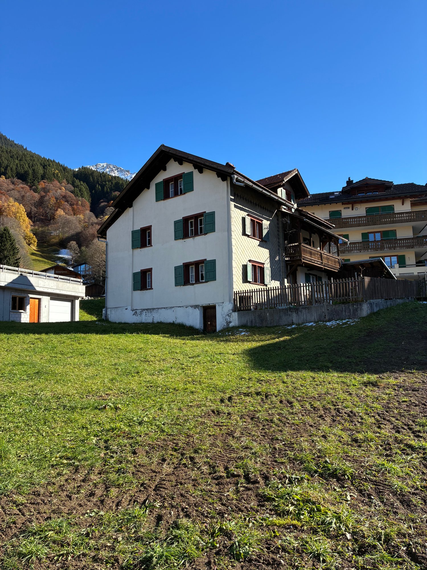 white house with green windows, wooden balcony, located on a hill with a mountain in the background