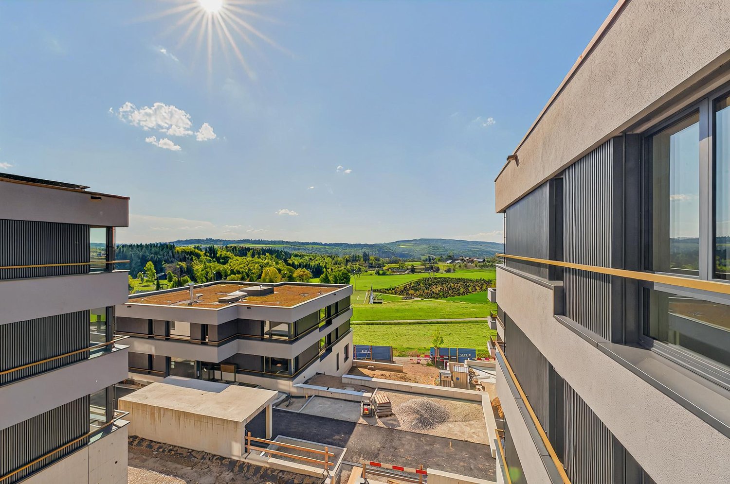 Modern multi-story building complex, overlooking a large green field and surrounding landscape, concrete structure, glass windows, roof with a terrace