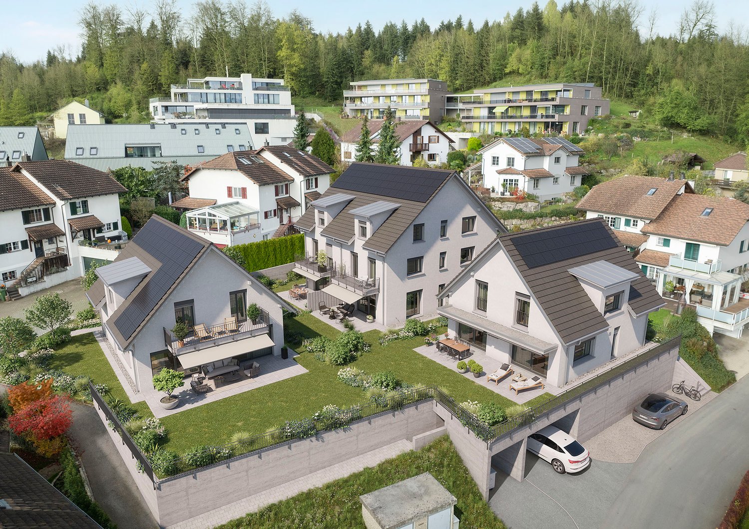 The image shows a residential neighborhood with a mix of multi-story apartment buildings and smaller houses. The buildings have balconies and terraces, and there is a parking area visible. The neighborhood is surrounded by a forested area, creating a scen