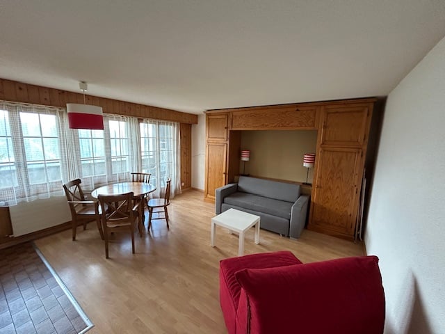 Spacious living room with wooden floors, large windows, dining table, sofa, and a balcony or terrace visible.