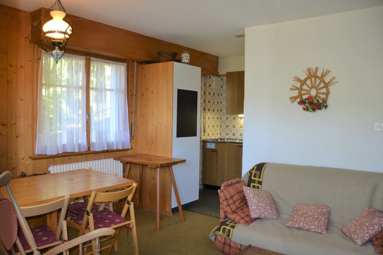 wooden walls, window with white curtains, small kitchen area, wooden dining table with chairs, couch with floral pillows, lamp