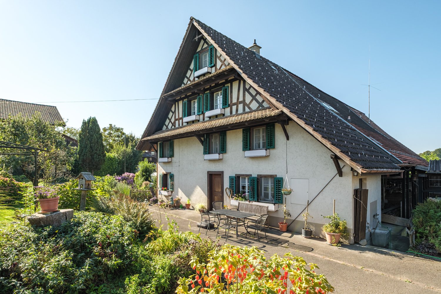 2 story house, white exterior, green shutters, brown roof, garden, seating area, table and chairs