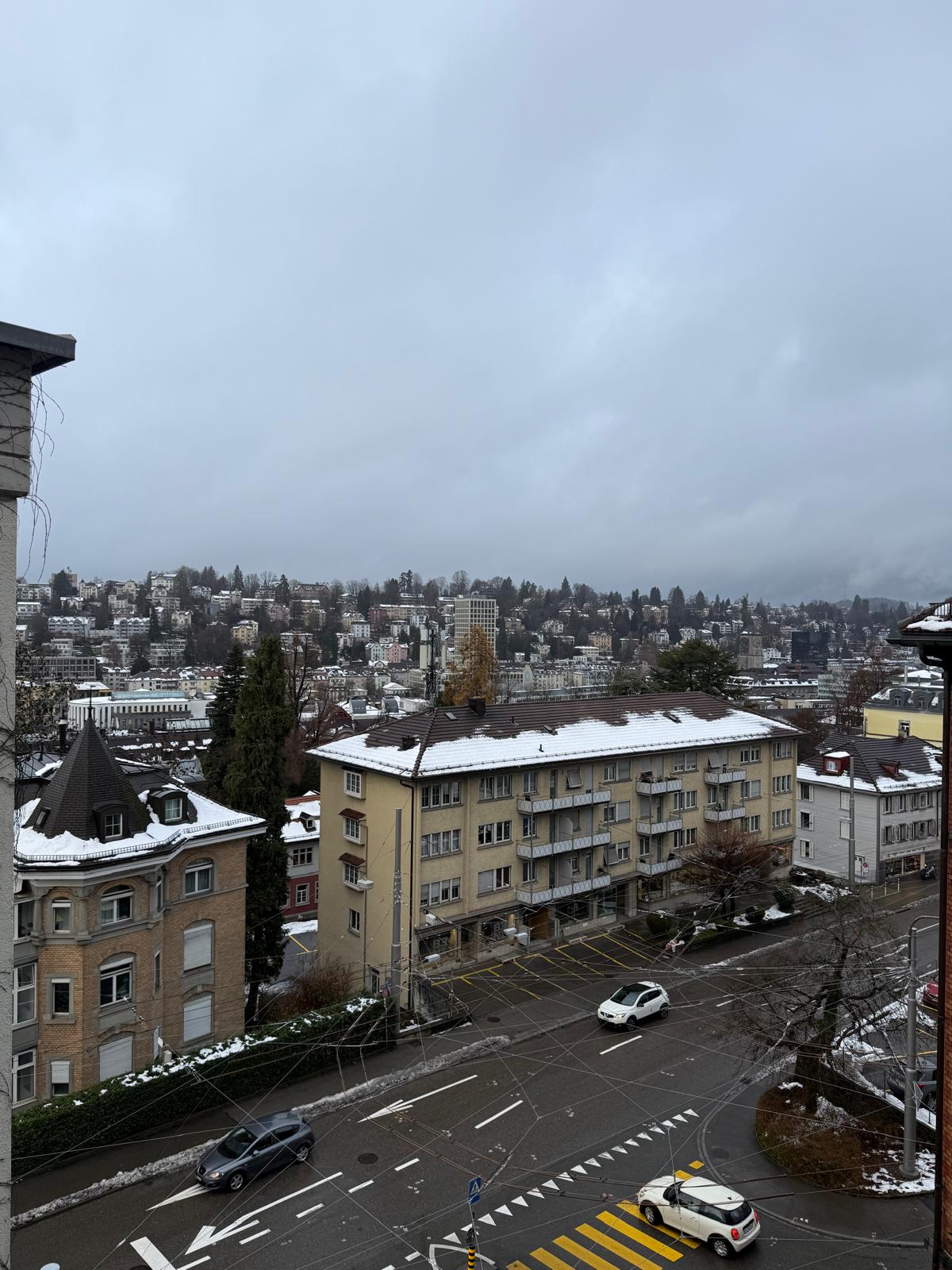 Urban landscape featuring a multi-story building with snow on the roof, neighboring structures, trees, parked cars on the road, pedestrian crossing with street lights, power lines, and a snowy street