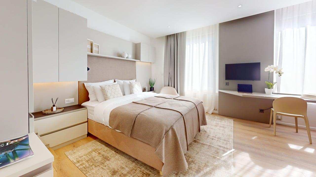 modern bedroom with wooden floor, gray and white walls, king-size bed, beige carpet, white nightstands, desk with laptop and TV, white curtains, and chairs