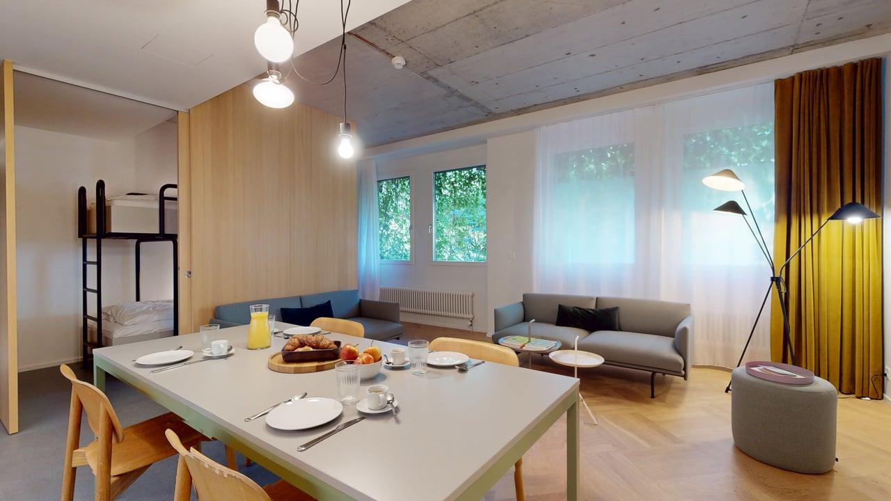 large dining area, white table, dining chairs, a white ottoman, gray couch, a small end table, blue sofa, a small table, lamps, white curtains, a yellow blanket, bunk beds, a radiator, a white door