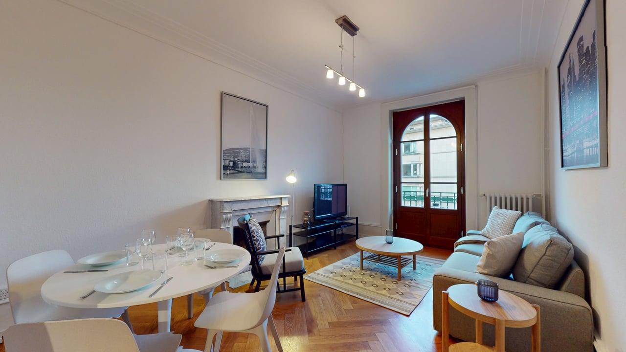 white painted walls, white hardwood floors, a fireplace, a large glass door with a window, a round wooden coffee table, a rug, a grey sectional sofa, a dining area with a round white table and chairs, a TV with a TV stand, a side table with a bowl, a lamp