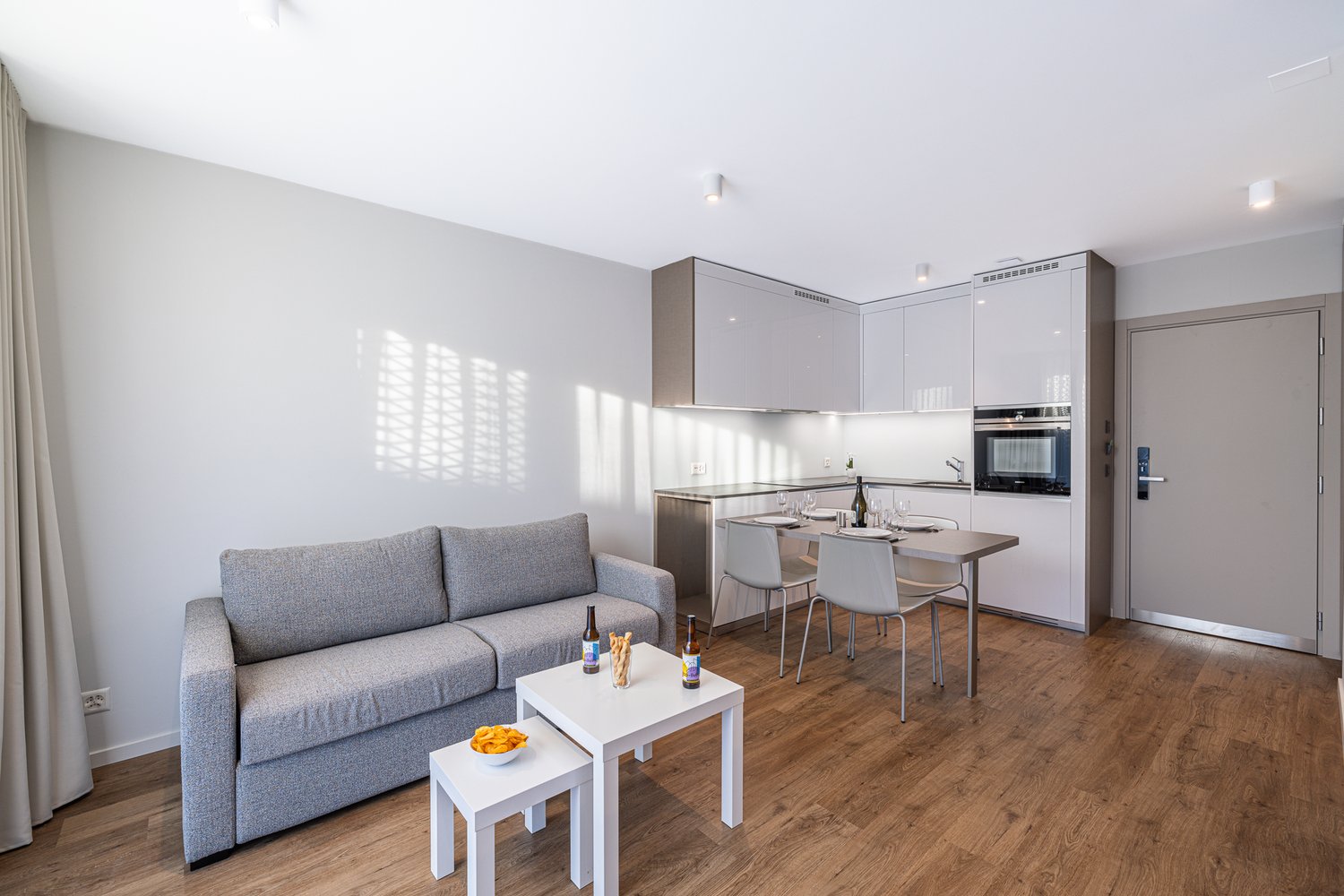 Open space living room with kitchen, dining area, and seating area with a gray couch, white tables, wooden floor, white kitchen cabinets, and stainless steel countertops.
