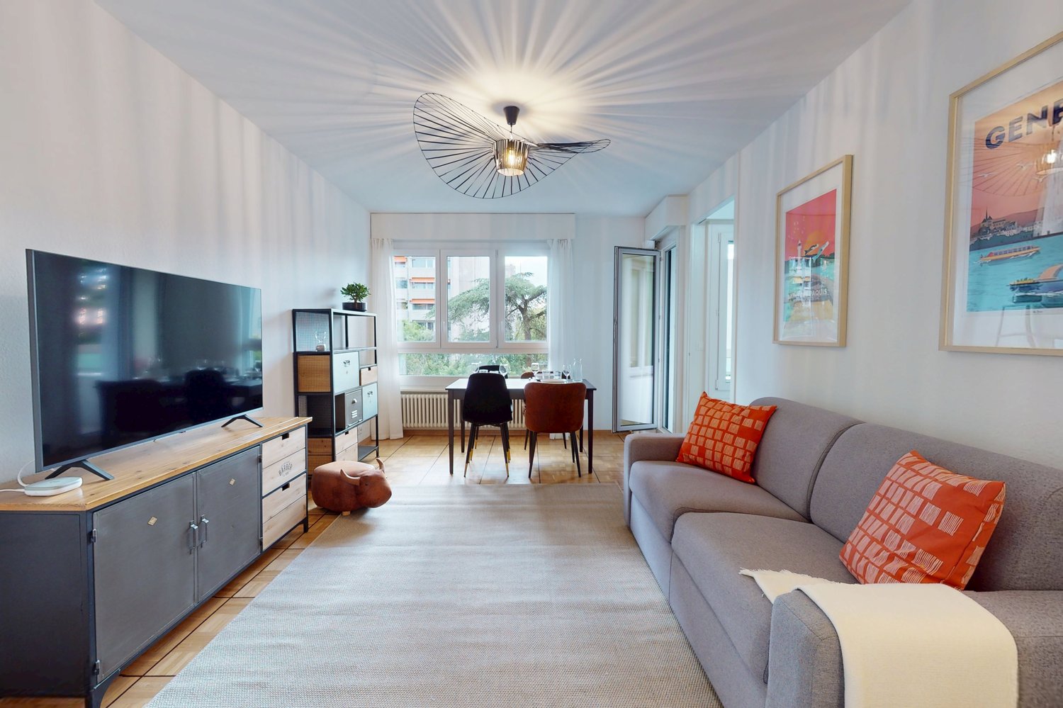 Living room with grey sofa, flatscreen TV, dining table, oak parquet floor, ceiling lamp, pictures on the wall, radiator, open door to another room.