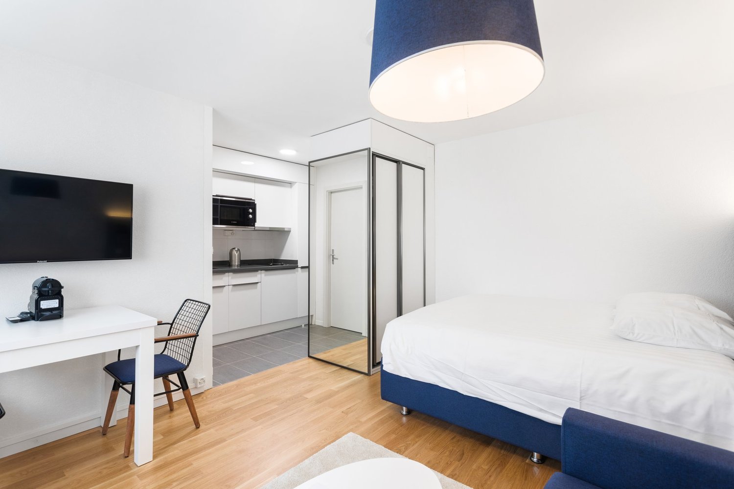 The image shows a modern and minimalist studio apartment. The room features a white and blue color scheme, with a large bed, a desk with a chair, and a kitchenette area with a microwave and other appliances. The room has hardwood floors and a large window