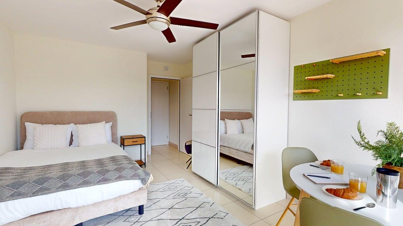 Bedroom with a bed, pillows, nightstand, ceiling fan, a closet with a mirror, a dining table with chairs, a pot plant, breakfast items