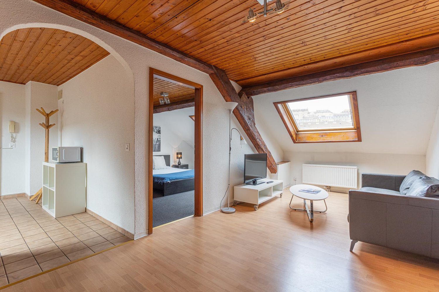 modern apartment with wooden floors, wooden beam ceiling, cozy living room with couch, TV, desk, bedroom with bed, microwave on stand, bedroom with skylight