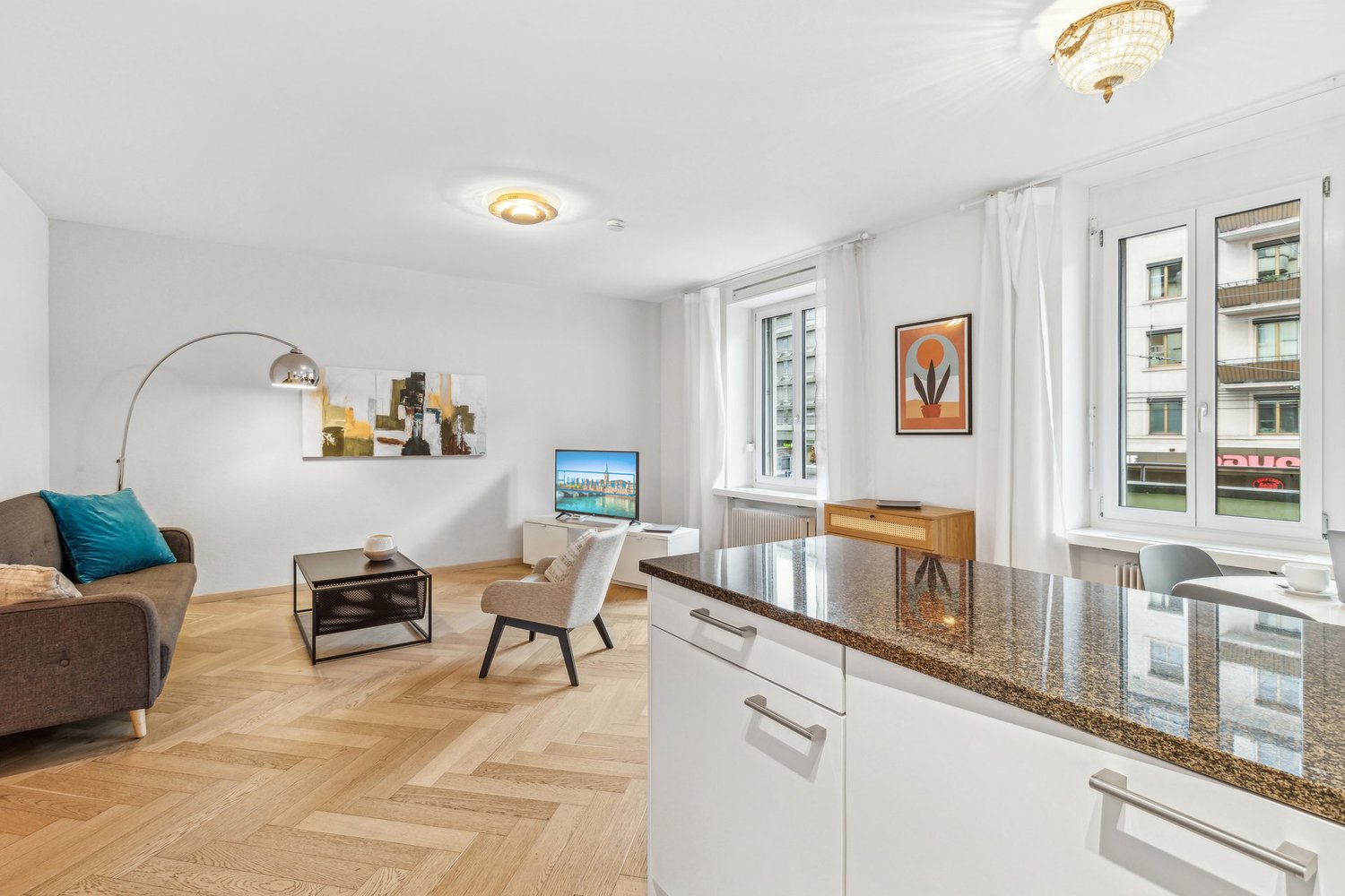 open living space with kitchen, large couch with cushions, armchair, coffee table, tv stand with flat screen tv, floor lamp, wooden parquet flooring, kitchen island with granite countertop, dining area with dining table and chairs, picture frames on the w