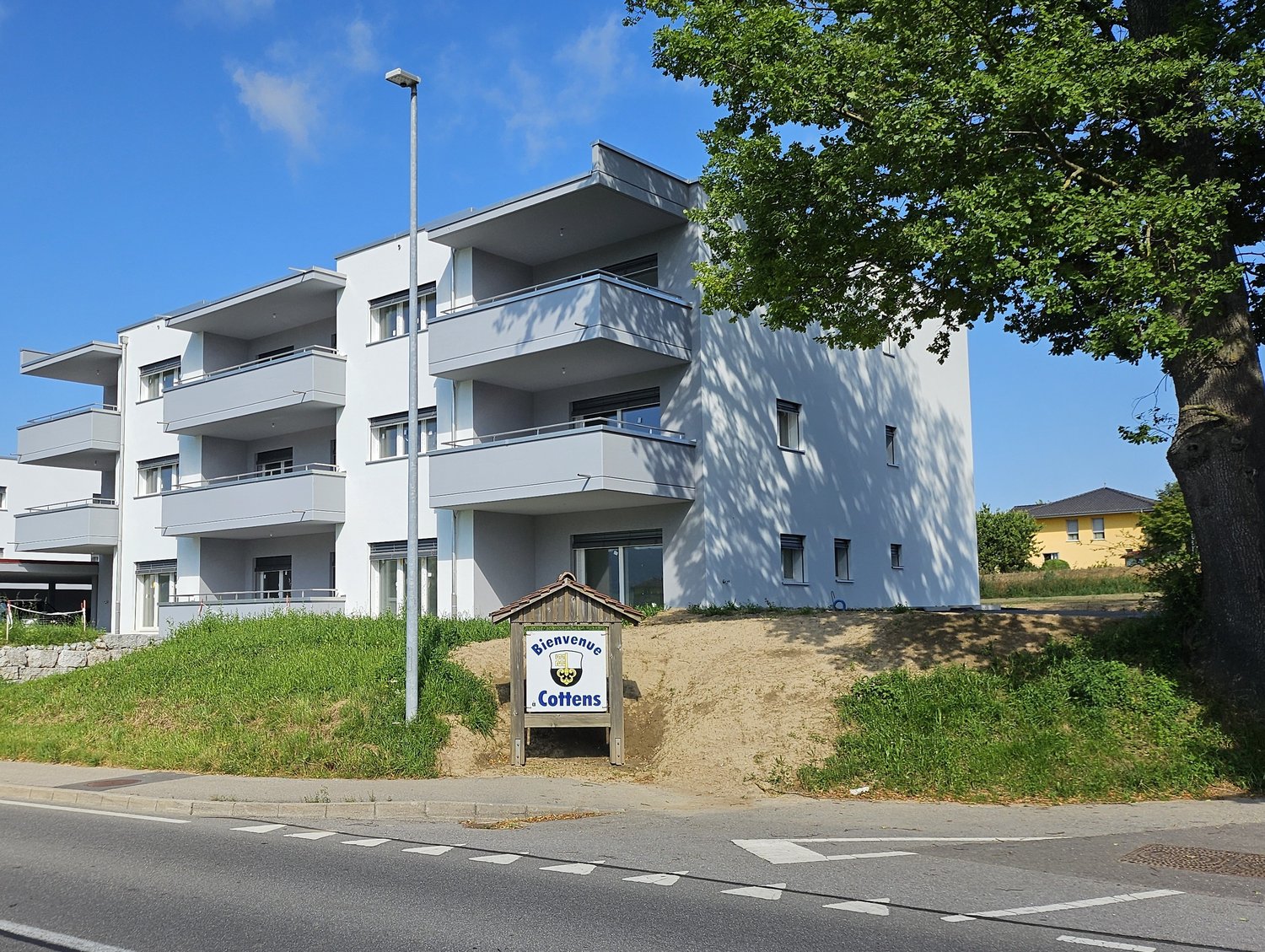 3-story apartment building with white exterior, balconies, and trees surrounding the property. There is a sign in the foreground that says 'Bienvenue Cottens'.