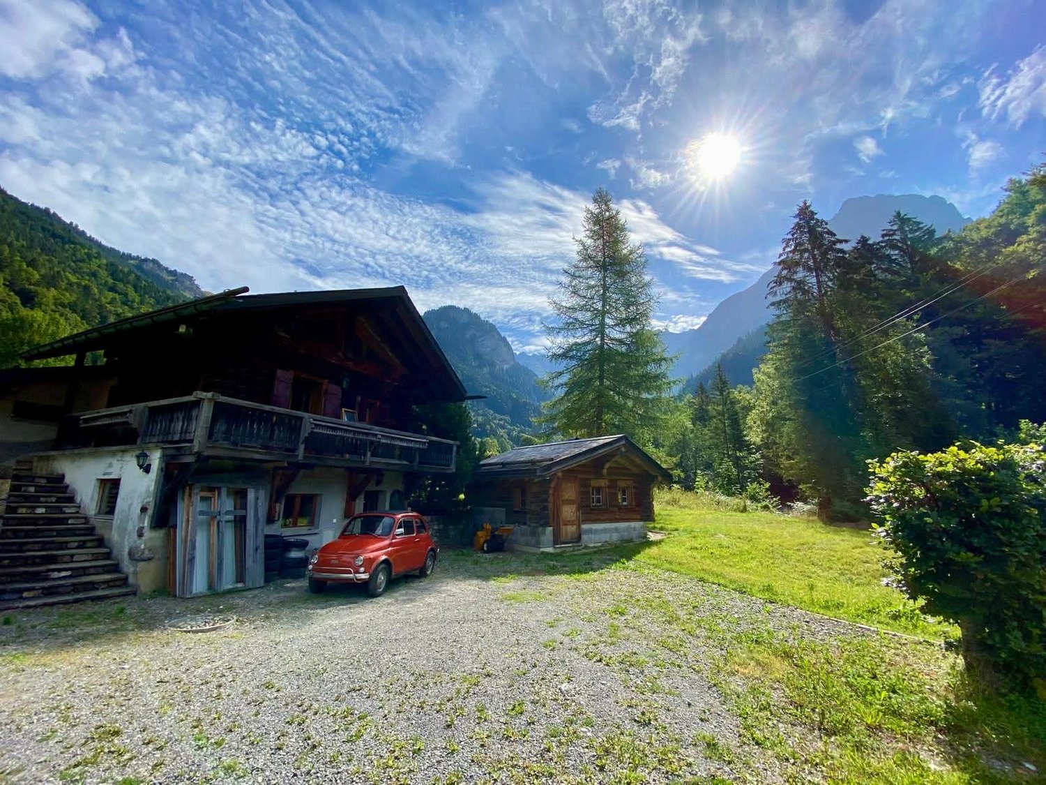 2 story wooden house, gravel driveway, steps leading to porch, red car, small wooden cabin, sunny weather, mountains in the background