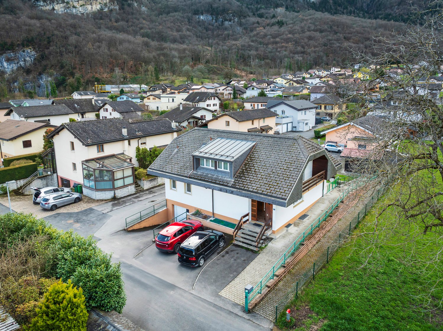 Aerial view of a small town or village nestled in a mountainous, forested landscape. The image shows a cluster of residential buildings with tiled roofs, some with balconies or terraces. There are several parked cars visible in the foreground, indicating