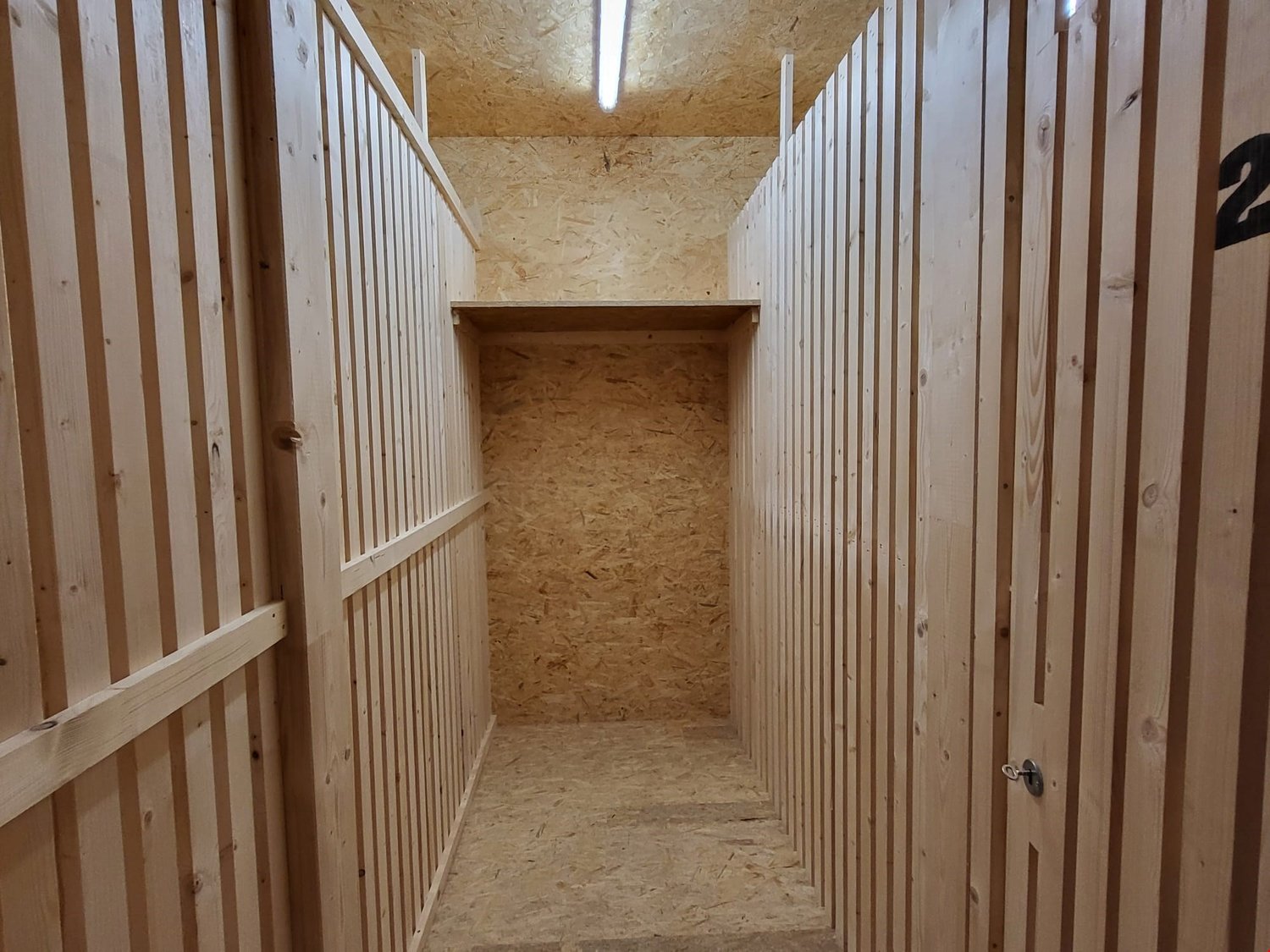 The image shows a long, narrow storage unit with wooden walls and shelves. The unit appears to be divided into individual compartments or lockers, each with a door. The walls and ceiling are made of oriented strand board (OSB) material, giving it a rustic