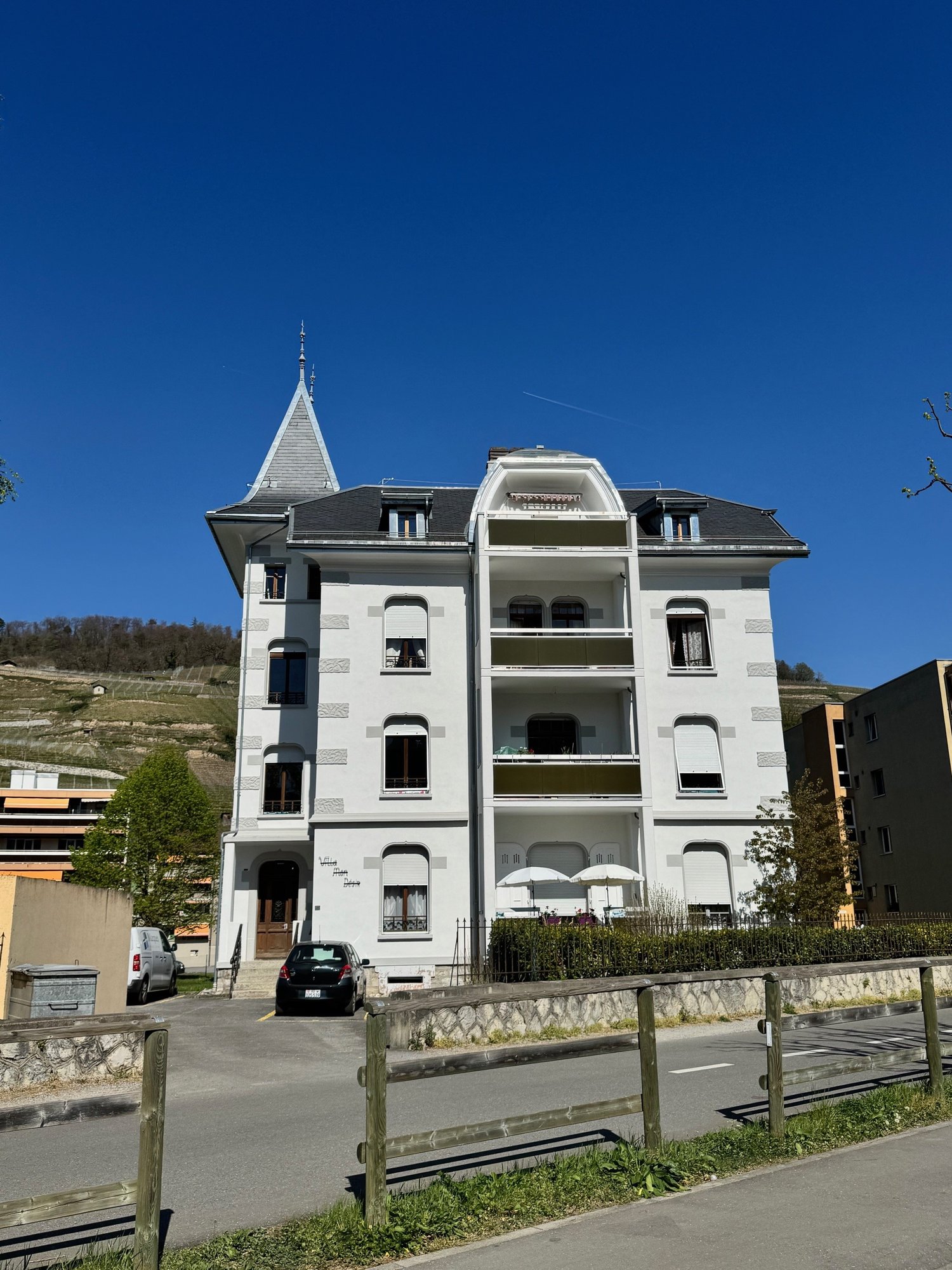 4 story house, white, multiple balconies, gray roof, black car parked in front, trees and plants around, mountains in the background