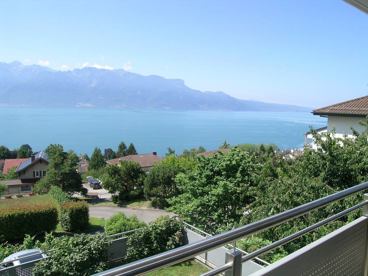 Balcony overlooking a serene lake and mountains in the distance with several houses and greenery.