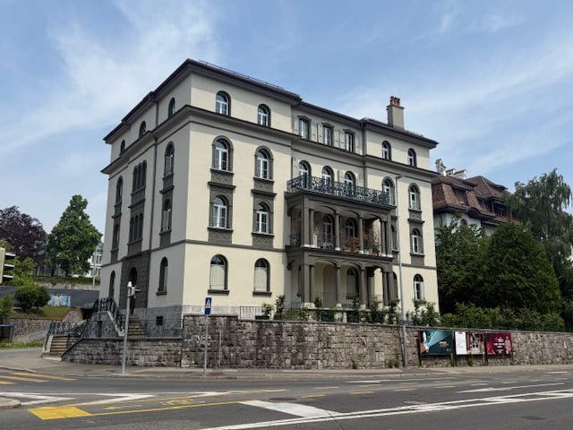 A large, multi-story building with a classical architectural style, featuring arched windows, balconies, and a stone facade. The building appears to be located in an urban setting, with a road and crosswalk visible in the foreground.