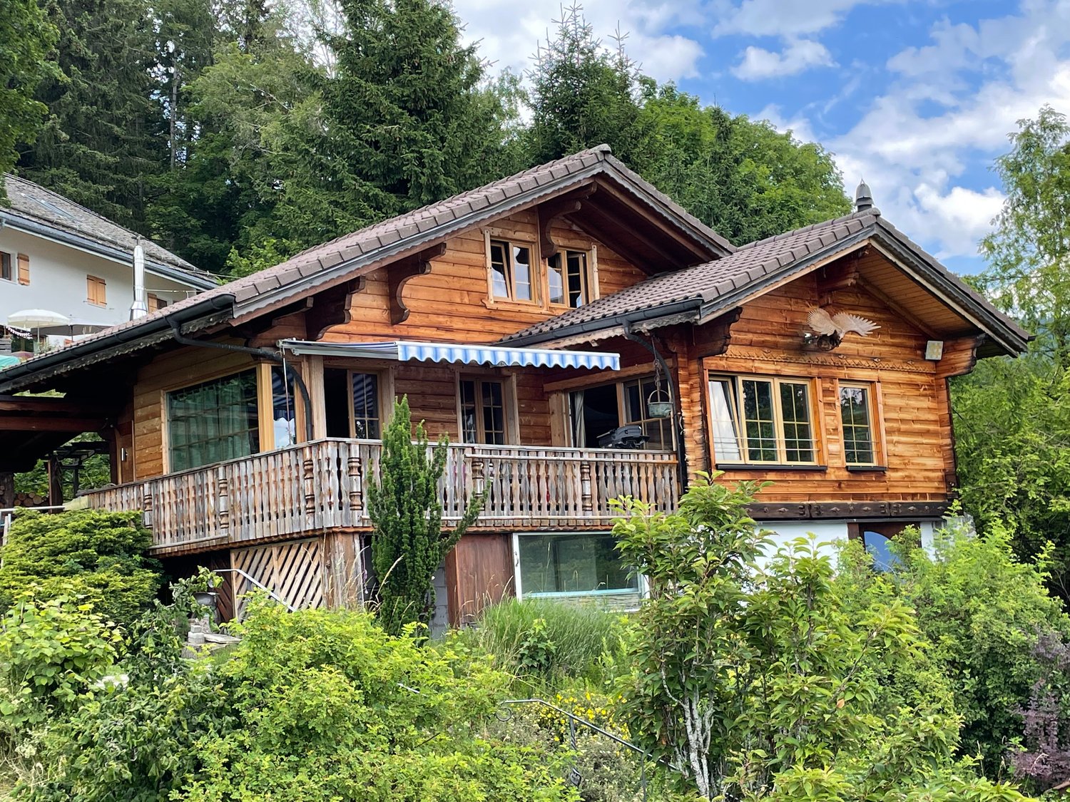 Two-story wooden chalet-style house with a balcony, surrounded by lush greenery and trees
