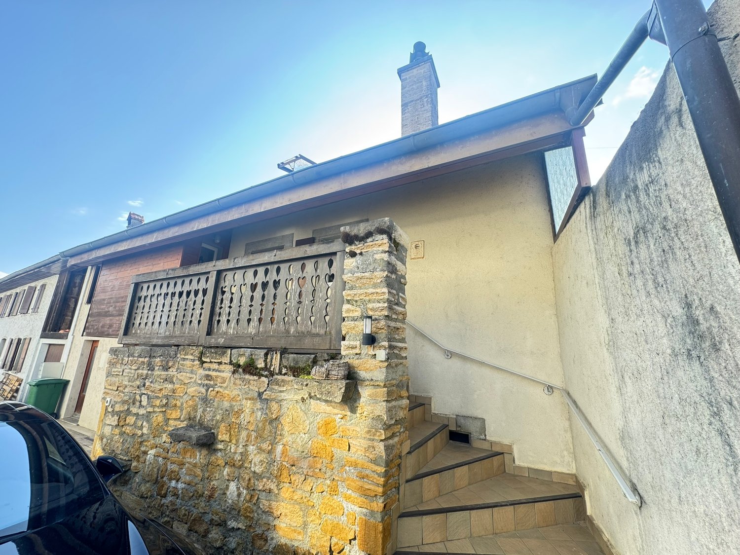 Two-story house with a raised ground floor, stone and stucco exterior, decorative balcony railing, and a chimney on the roof.