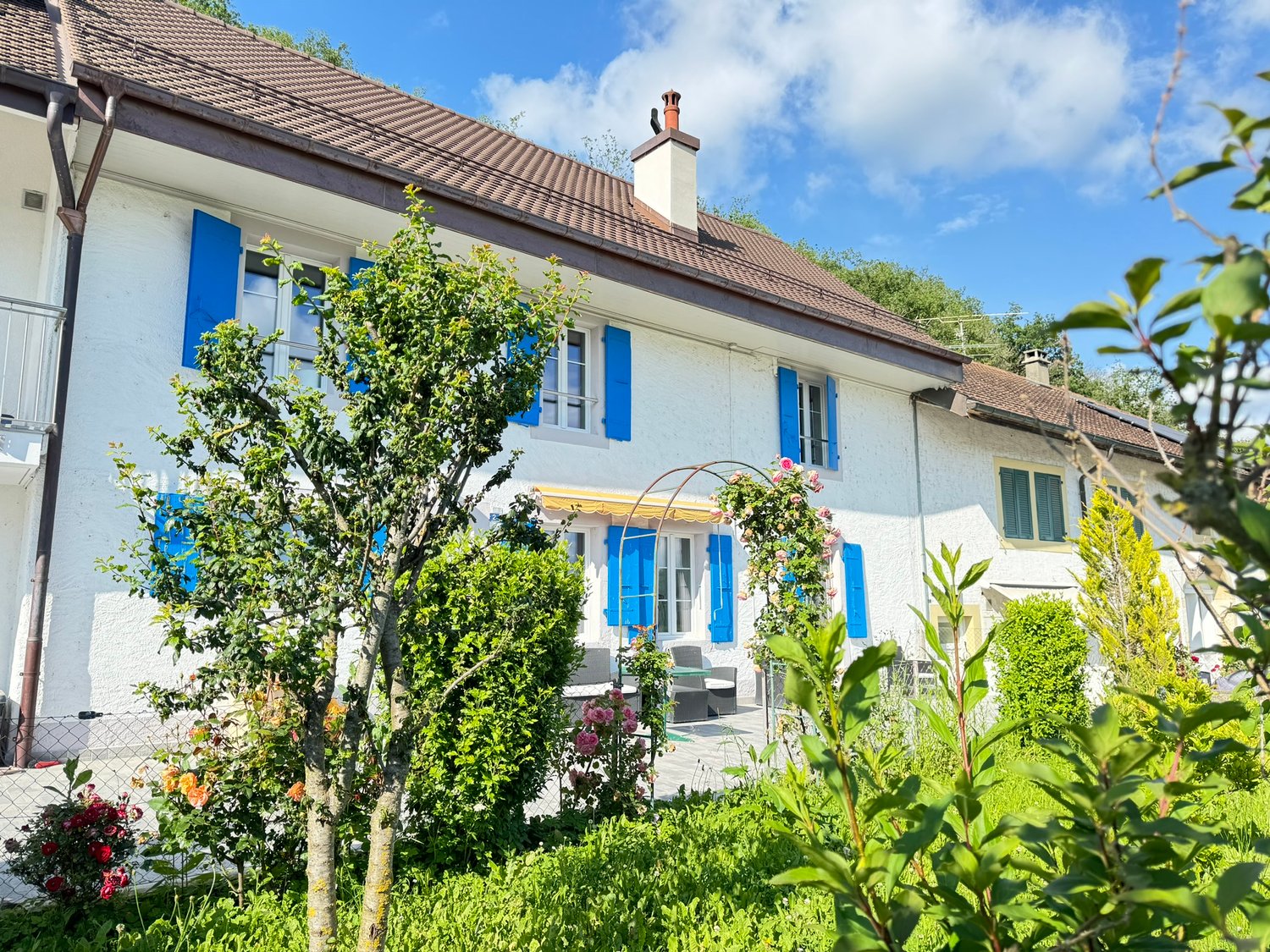2 story house, brown roof, white walls, blue shutters, outdoor seating, garden