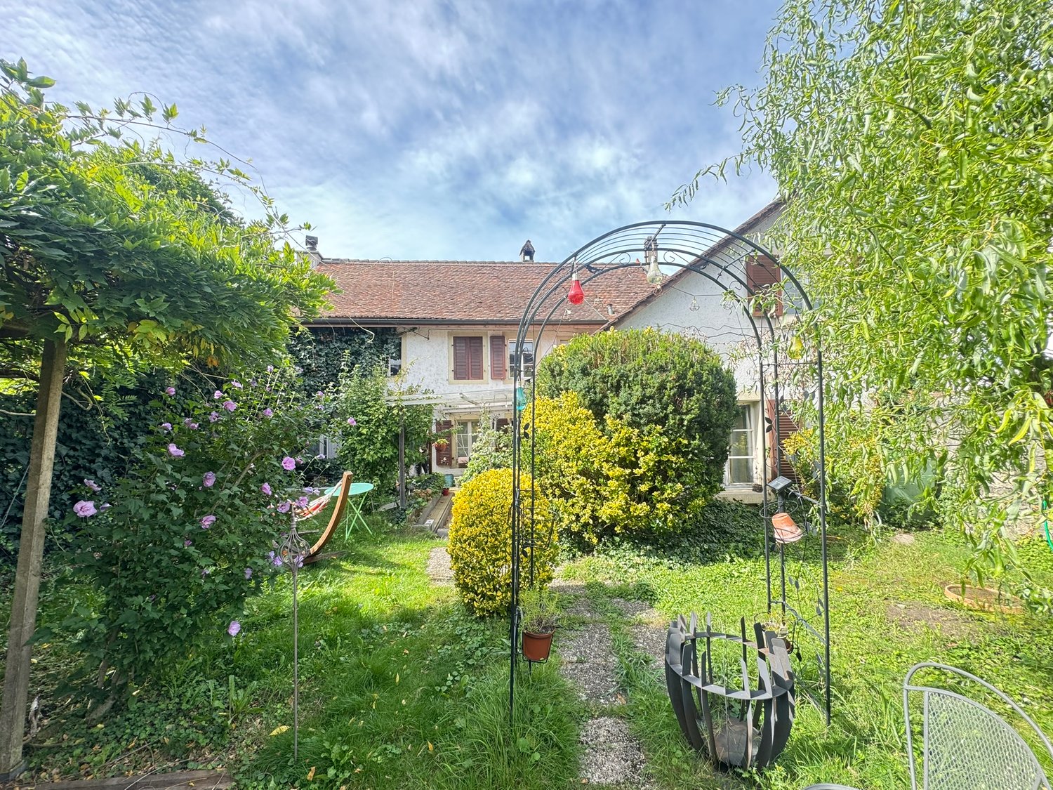 well-maintained garden with various plants, trees, shrubs, pathway, metal arch, patio set, metal trellis, and a house in the background