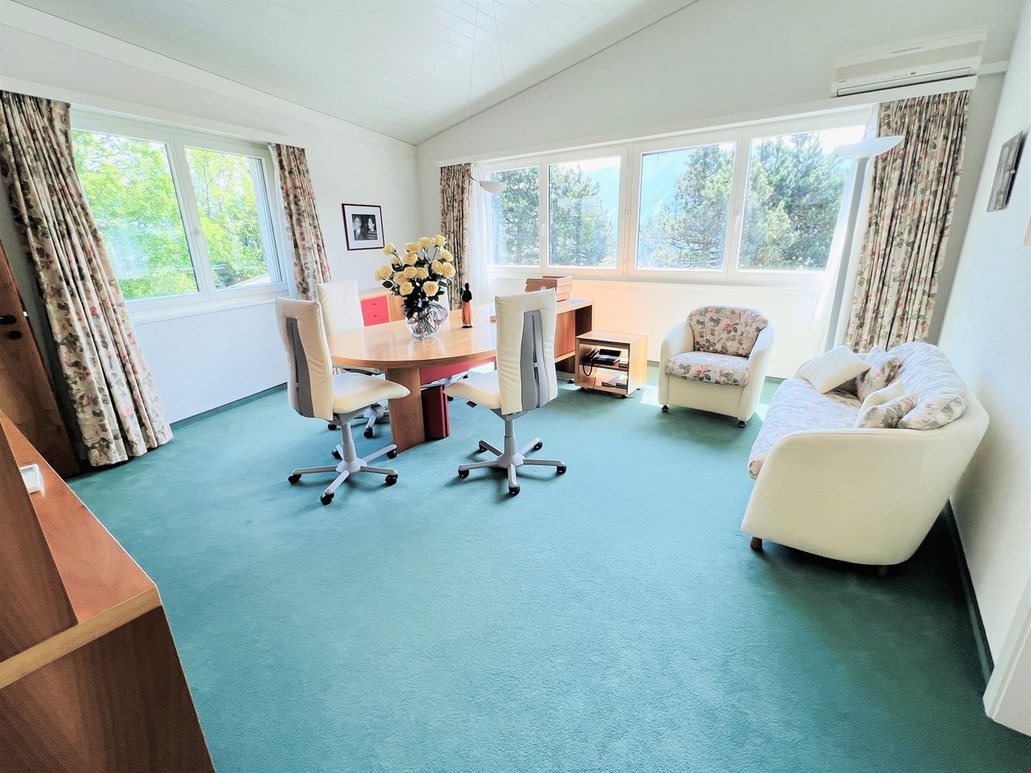 Spacious home office with large windows, wooden desk, office chairs, floral armchair, and a sofa. The room has a blue carpeted floor and appears to have access to a balcony or terrace.