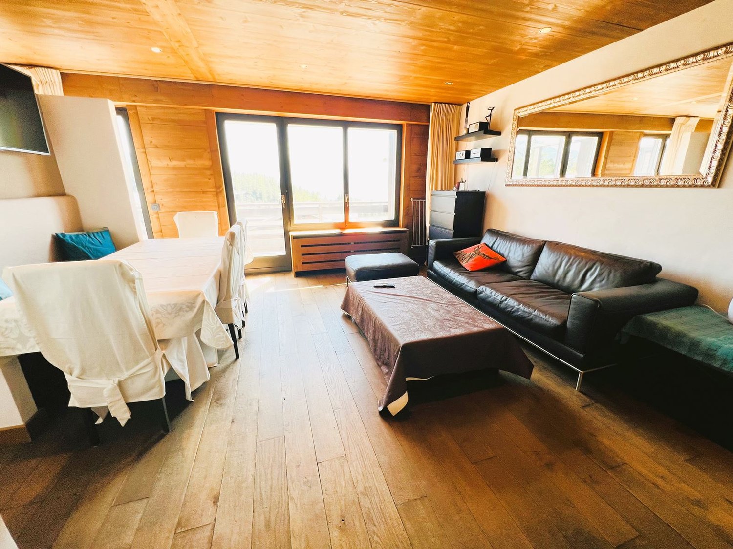 Spacious living room with wooden walls and ceiling, large windows, leather sofa, coffee table, and a balcony or terrace visible.